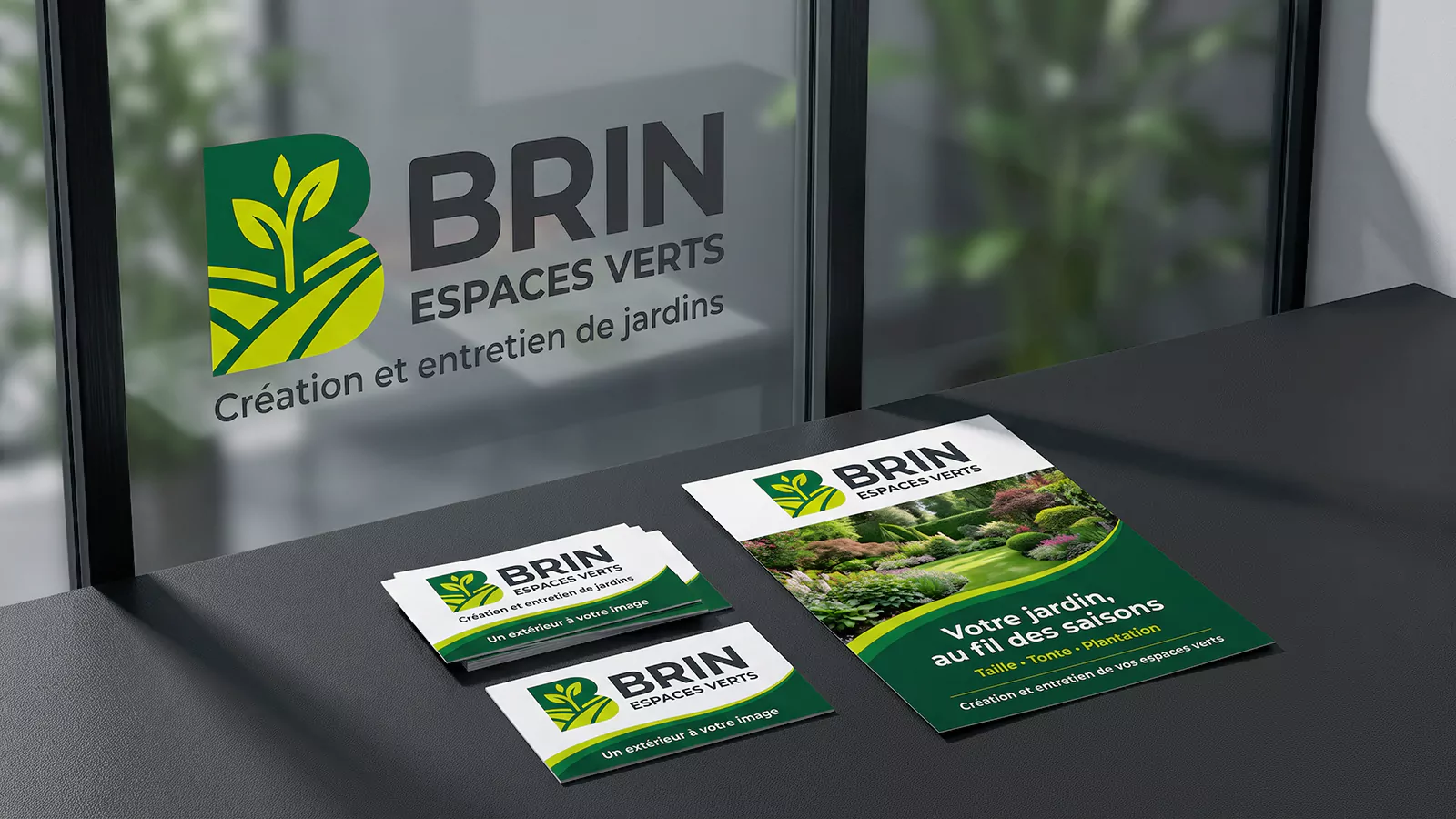 Logo BRIN Espaces verts en lettrage adhésif sur une vitre, avec des cartes de visite et un flyer posés sur un plan de travail sombre.