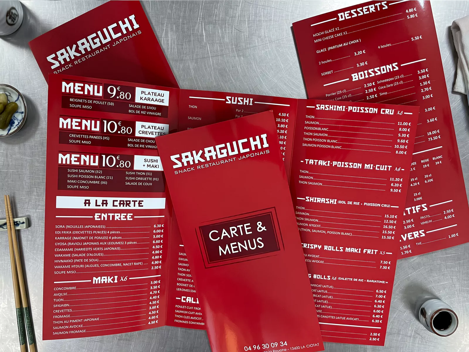 Cartes de menu rouges Sakaguchi, fermées et ouvertes sur leurs trois volets, présentées sur un plan de travail en acier.
