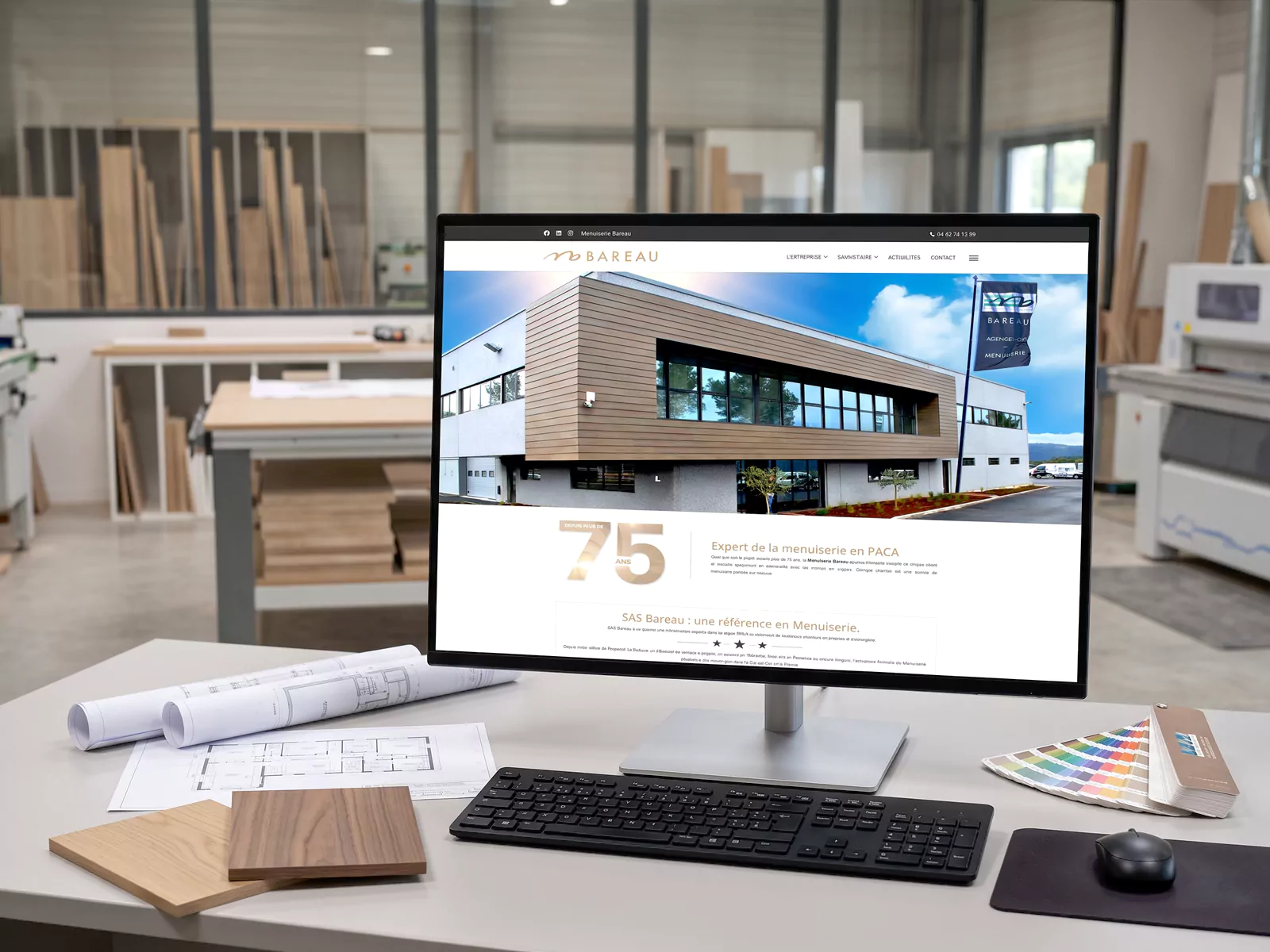 Page d’accueil du site de la Menuiserie Bareau affichée sur un écran dans un bureau d’étude, avec des plans et des échantillons de bois.